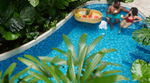 Casal relaxando em piscina cercada por vegetação tropical, desfrutando bebidas naturais ao lado de uma bandeja flutuante com frutas, em ambiente climatizado por bomba de calor.