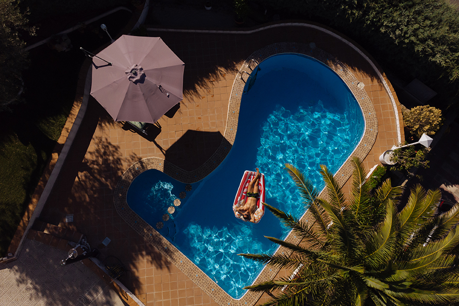 Vista aérea de uma piscina com mulher relaxando em boia inflável sob o sol, destacando o aproveitamento completo da área de lazer com conforto térmico garantido por bomba de calor.