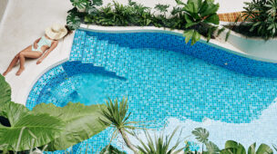 Mulher relaxando à beira de uma piscina azul com design orgânico e cercada por vegetação tropical, em ambiente climatizado com trocador de calor.