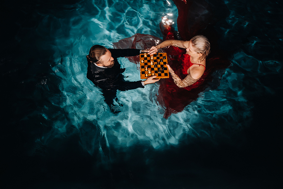 Casal elegante jogando xadrez dentro de uma piscina iluminada à noite, representando conforto térmico e experiências sofisticadas.