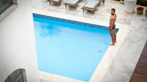 Homem ao lado de piscina residencial, observando a água em ambiente elegante com espreguiçadeiras, representando a importância do conforto térmico com bomba de calor.